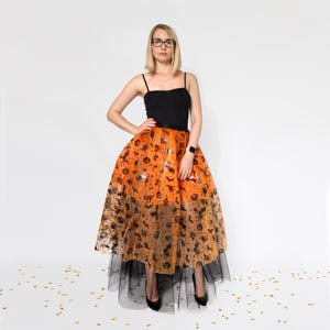 May include: A black camisole top paired with an orange Halloween-themed skirt. The skirt features a pattern of pumpkins, bats, and stars. The skirt has a black tulle underskirt. The person is wearing black heels.