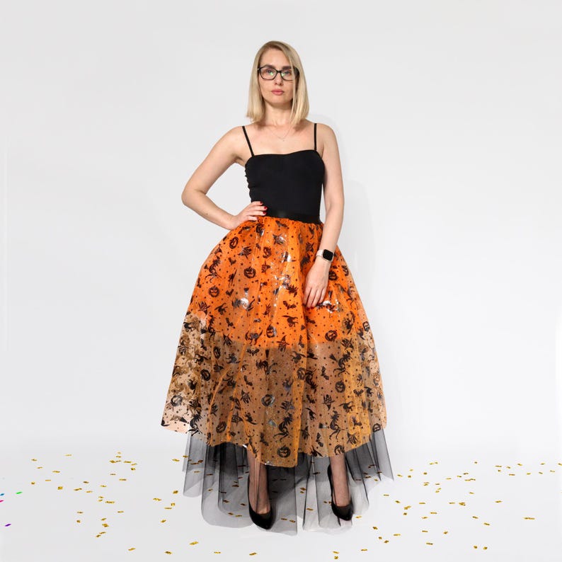 May include: A black camisole top paired with an orange Halloween-themed skirt. The skirt features a pattern of pumpkins, bats, and stars. The skirt has a black tulle underskirt. The person is wearing black heels.