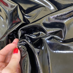 Op de afbeelding: Close-up van een glanzende, zwarte stof met een reflecterend oppervlak. Het materiaal is gevouwen en gekreukt, waardoor highlights en schaduwen ontstaan. De stof ziet er glad uit en heeft een glanzende afwerking. Een hand met rode nagellak raakt de stof aan.