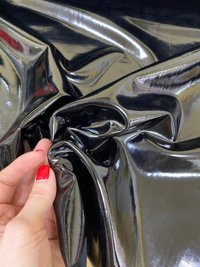 Op de afbeelding: Close-up van een glanzende, zwarte stof met een reflecterend oppervlak. Het materiaal is gevouwen en gekreukt, waardoor highlights en schaduwen ontstaan. De stof ziet er glad uit en heeft een glanzende afwerking. Een hand met rode nagellak raakt de stof aan.