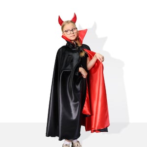 May include: A child dressed as a devil for Halloween. They are wearing a black cape with a red lining and red devil horns. The child is looking to the side and has a serious expression on their face.