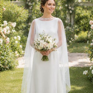 May include: A white wedding dress with a sheer cape and a floral neckline. The bride holds a bouquet of white and cream-colored flowers. The background features a garden with white flowers and greenery.