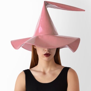 May include: A pink witch hat with a wide brim and a tall, pointed crown. The hat has a glossy finish and a curved brim.