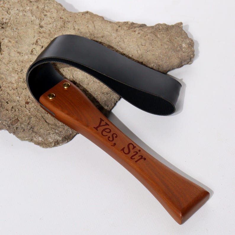 May include: A wooden paddle with a black leather strap. The paddle is engraved with the words "Yes, Sir". The wood is a rich brown color, and the leather is black.