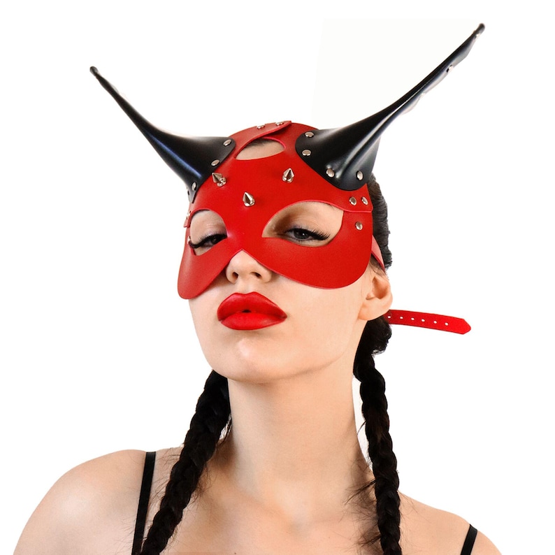 Puede incluir: Una m&aacute;scara de cuero roja con cuernos negros y tachuelas plateadas. La m&aacute;scara tiene un recorte para los ojos y una peque&ntilde;a abertura para la nariz. La m&aacute;scara la lleva puesta una persona con trenzas negras largas.