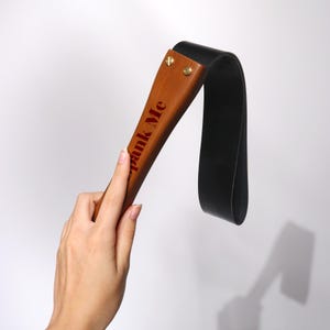 May include: A wooden paddle with the text "Spank Me" in red lettering. The paddle has a black leather strap attached with brass rivets. The item is held by a hand.