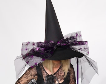 May include: Black witch hat with a tall, pointed crown. The hat features a wide brim adorned with a large bow made of sheer black fabric with purple spider accents. Black tulle and purple ribbons cascade from the brim.