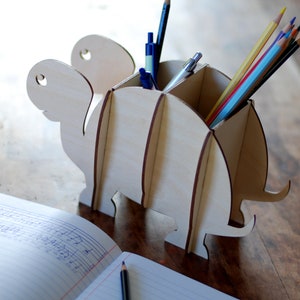 May include: Wooden turtle-shaped desk organizer with four compartments for pens and pencils. The organizer is made of light brown wood and has a natural finish. It is filled with colorful pencils and pens.