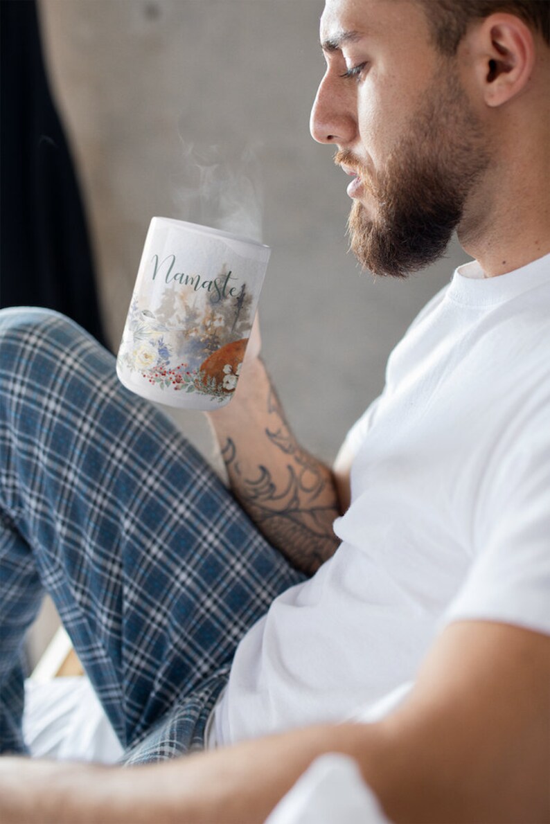 May include: A white ceramic mug with a watercolor design of a forest scene and the word "Namaste" printed on the front. The mug is filled with a hot beverage and steam is rising from the top.