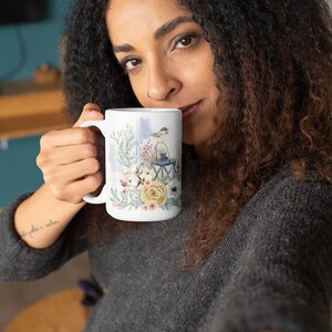 May include: A white ceramic mug with a winter scene featuring a bird perched on a lantern, surrounded by flowers and greenery. The mug has a handle and is being held by a person.