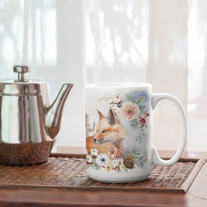May include: White ceramic mug with a watercolor design of a fox surrounded by flowers and pine cones. The fox is brown and orange with a white chest. The flowers are white, pink, and yellow.