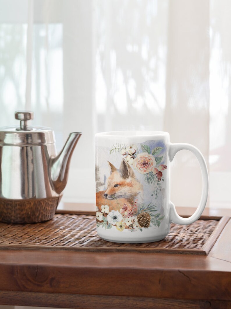 May include: White ceramic mug with a watercolor design of a fox surrounded by flowers and pine cones. The fox is brown and orange with a white chest. The flowers are white, pink, and yellow.