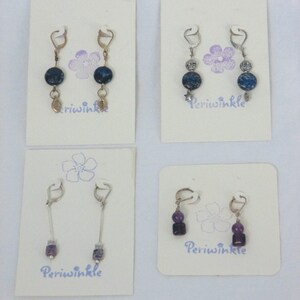 Puede incluir: Cuatro pares de pendientes en tarjetas blancas con la palabra "Periwinkle" impresa en cada tarjeta. Los pendientes están hechos de metal plateado y dorado y presentan piedras azules y moradas.