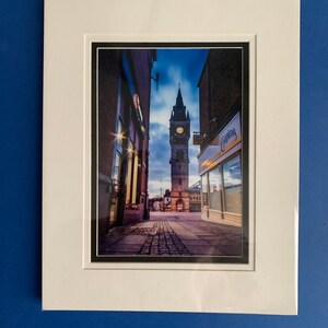 May include: A view down a narrow street in a town, with a clock tower in the distance. The street is lined with buildings, and the sky is a deep blue. The clock tower is lit up, and the street is empty.