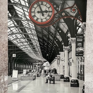 May include: A black and white photo of a train station interior with a large red clock hanging from the ceiling. The clock has black Roman numerals and hands. The station is empty except for a few people walking around. There are signs for "AZUMA", "HITACHI POWER", and "LNER PRIDE".