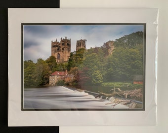 May include: A photograph of a cathedral with a river in the foreground. The cathedral is made of stone and has a tall spire. The river is flowing over a small dam. The sky is cloudy and the trees are green.