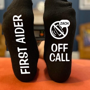 May include: A pair of black socks with white text. One sock says "FIRST AIDER" vertically. The other sock features a graphic of a phone with a line through it and the word "ZACH" in a speech bubble, with the text "OFF CALL" below.