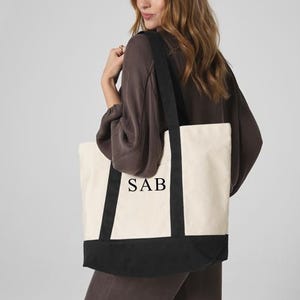 Puede incluir: Bolso tote de lona color crema con detalles y asas negras. La parte inferior de la bolsa es negra y el texto "SAB" está impreso en negro en la parte delantera. El bolso se lleva al hombro.