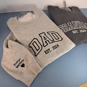 May include: Two gray sweatshirts with black text. One sweatshirt says "DAD EST. 2024" and the other says "GRANDPA EST. 2024". The sweatshirt with "DAD" has the names "Evelyn Matilda" and a heart on the sleeve.