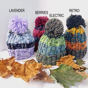 May include: Four colorful knit beanies with pom-poms. The beanies are labeled "LAVENDER", "BERRIES", "ELECTRIC", and "RETRO". The beanies are in shades of purple, red, green, blue, yellow, and orange. Dried leaves are scattered in front of the beanies.