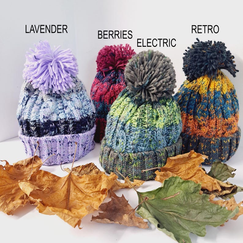May include: Four colorful knit beanies with pom-poms. The beanies are labeled "LAVENDER", "BERRIES", "ELECTRIC", and "RETRO". The beanies are in shades of purple, red, green, blue, yellow, and orange. Dried leaves are scattered in front of the beanies.