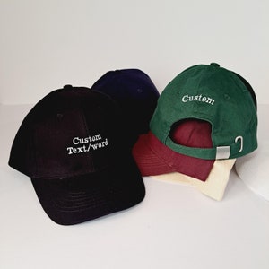 Op de afbeelding: Drie baseballpetten met verstelbare bandjes. Een zwarte pet met witte borduurwerk dat "Custom Text/word" zegt, een groene pet met witte borduurwerk dat "Custom" zegt, en een donkerblauwe pet.