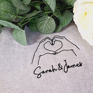 May include: Gray sweatshirt with a black embroidered design of two hands forming a heart shape with the text "Sarah & James" below.