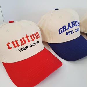 May include: Cream-coloured baseball caps with red and blue visors. One cap has "custom" embroidered in red, and "YOUR DESIGN" in black. Another cap has "GRANDAD EST. 2024" embroidered in blue. The caps are a classic style.