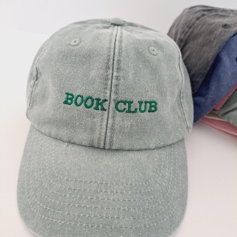 May include: A light grey baseball cap with a green embroidered logo that says "BOOK CLUB".