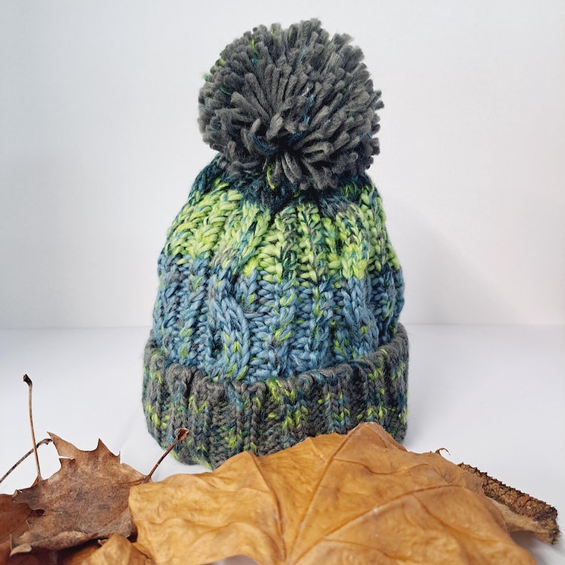 May include: A hand-knitted winter hat with a large gray pom-pom. The hat features a cable knit design in shades of blue, green, and gray. The brim is a darker gray. The hat is displayed on a white surface with dried leaves.