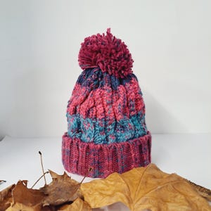 May include: A hand-knitted winter hat with a cable knit design. The hat features a color scheme of pink, teal, and navy blue, topped with a large, matching pom-pom. The brim is a solid pink color.
