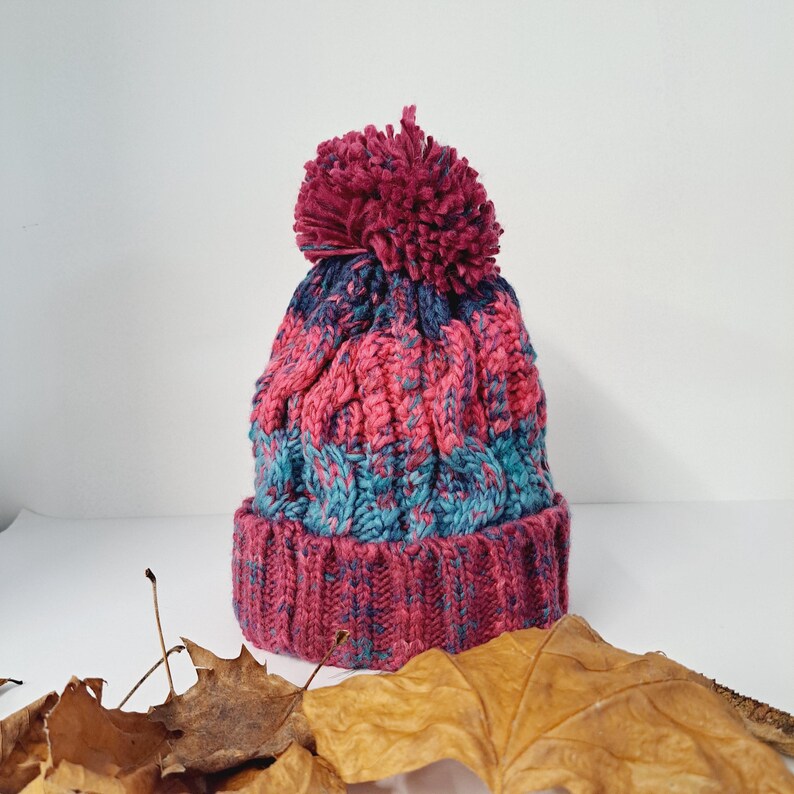 May include: A hand-knitted winter hat with a cable knit design. The hat features a color scheme of pink, teal, and navy blue, topped with a large, matching pom-pom. The brim is a solid pink color.