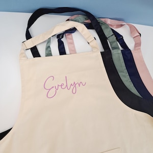 May include: A beige canvas apron with adjustable straps. The apron has a large pocket on the bottom and the name "Evelyn" is embroidered in pink on the front.