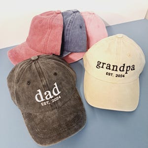 May include: Four baseball caps in different colours with embroidered text. The caps are black, beige, pink, and blue. The text on the caps reads "dad est. 2024", "grandpa est. 2024", and "est. 2024".