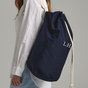 以下が含まれることがあります： 白のロープストラップと白で印刷されたイニシャル「LH」が付いた、ネイビーブルーの巾着袋。バッグは柔らかく、わずかに光沢のある素材で作られています。バッグは肩にかけられています。