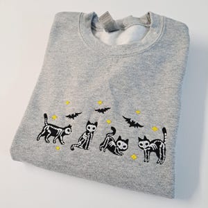 May include: A gray sweatshirt with a black and white embroidered design of four skeleton cats and bats. The cats are in different poses and the bats are flying. There are small yellow stars scattered around the design.
