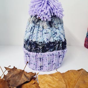 May include: A knitted winter hat with a large lavender pom-pom. The hat features a ribbed brim in a light purple color, transitioning to a textured pattern in shades of blue, gray, and navy. The hat is designed for cold weather.