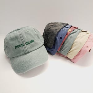 Op de afbeelding: Een stapel van acht baseballpetten in verschillende kleuren, waaronder grijs, blauw, rood, beige en roze. Eén pet is groen en heeft de tekst "BOOK CLUB" geborduurd op de voorkant.