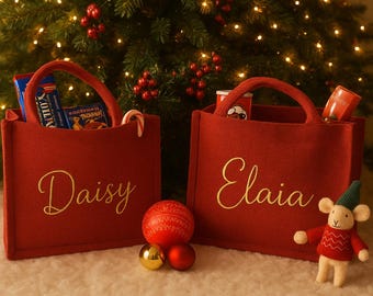 Könnte beinhalten: Zwei rote Tragetaschen mit goldgestickten Namen, "Daisy" und "Elaia". Jede Tasche enthält Snacks und einen Zuckerstange. Eine kleine Mäusefigur in einem roten Pullover und einer grünen Mütze steht neben den Taschen. Weihnachtsbaum im Hintergrund.