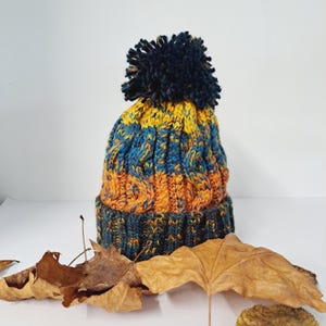May include: A hand-knitted beanie hat with a dark navy blue pom-pom. The hat features a cable knit design with bands of yellow, blue, and orange. The hat is displayed with dried leaves.