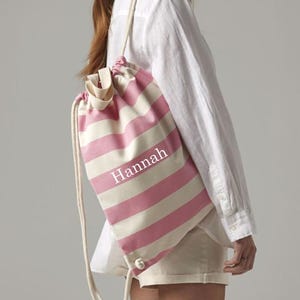 Puede incluir: Una mochila con cordón a rayas rosas y blancas con el nombre "Hannah" impreso en la parte delantera. La bolsa tiene un cordón de color crema y está hecha de un material similar a la lona.