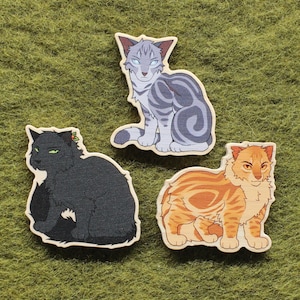 May include: Three wooden cat cutouts. One is gray with dark gray stripes, another is black, and the third is orange with brown stripes. The cats are on a green textured surface.