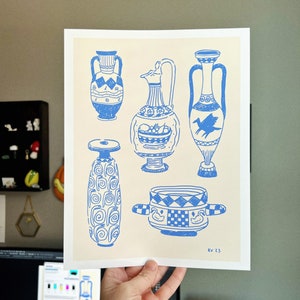 May include: A print of five blue and white illustrations of ancient Greek pottery. The illustrations include a pitcher, a vase, a bowl, and two amphorae. The print is on a cream-colored background and includes the text "KV 23".