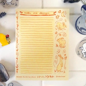 May include: A notepad with the words "notes and things" in red script, decorated with rabbit illustrations and floral accents. The notepad is surrounded by decorative stones, flowers, and a candle holder.