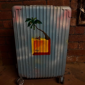 May include: A light blue hard-shell suitcase with vertical ridges, featuring a painted graphic of a palm tree on a yellow and red square block with the text esc. The suitcase has four wheels at the base and a soft, cloudy sky-like finish.