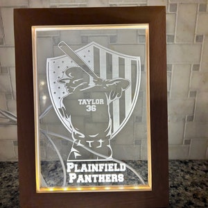 May include: A wooden framed illuminated sign with a white silhouette of a baseball player holding a bat in front of a shield with stars and stripes. The text "Taylor 36" is on the player's jersey and "Plainfield Panthers" is at the bottom of the sign.