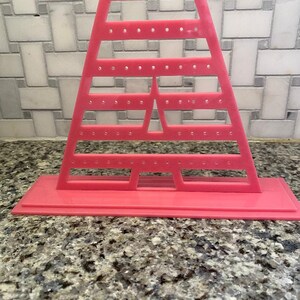 May include: Pink plastic earring holder with a triangular design and multiple holes for storing earrings. The holder has a rectangular base.