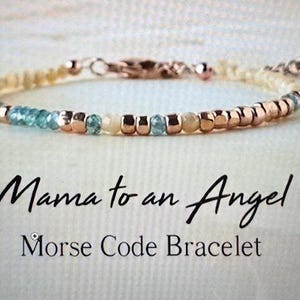 May include: A delicate beaded bracelet with a mix of gold, light blue, and cream-colored beads. The bracelet is inscribed with the words "Mama to an Angel" and "Morse Code Bracelet" in black script.