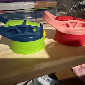 May include: Two colorful water bottle lids. One is navy blue and green, the other is red and pink. The lids have a flip-top design and are made of plastic. They are designed to fit on a water bottle.