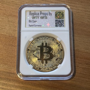Peut inclure: Une réplique de pièce Bitcoin dorée dans un étui en plastique transparent. La pièce présente le symbole Bitcoin et du texte sur le bord. L'étui porte une étiquette indiquant "Replica Props by UNITY GIFTS" et "Bit Coin Digital Currency."